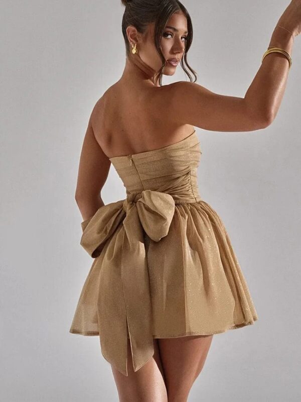 Woman in a gold strapless SEQUIN BOW CORSET DRESS with a large sequin bow on the back poses looking over her shoulder.