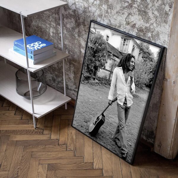 Black and white portrait photo of Bob Marley Roots with dreadlocks holding a guitar, leaning against a wall next to a bookshelf in a room with hardwood floors.
