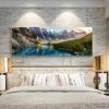 A modern bedroom features a large Landscape Wall Art of a mountainous lake above a neatly made bed with patterned pillows. The room has a neutral color scheme and metallic bedside lamps.