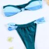 A two-piece bikini set, dubbed the EARTH ELEMENT TWO PIECE SWIMSUIT, features a twisted bandeau top and high-waisted bottoms in shades of blue and green. Displayed on a light surface, it is complemented by two small white flowers nearby.