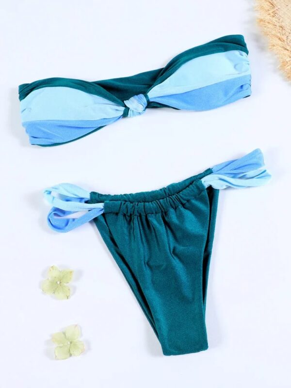 A two-piece bikini set, dubbed the EARTH ELEMENT TWO PIECE SWIMSUIT, features a twisted bandeau top and high-waisted bottoms in shades of blue and green. Displayed on a light surface, it is complemented by two small white flowers nearby.