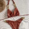 A TWO PIECE BRAZILIAN BIKINI SET with gold chain accents is laid flat on a light fabric surface. A brown beret with pearl decorations is positioned nearby.