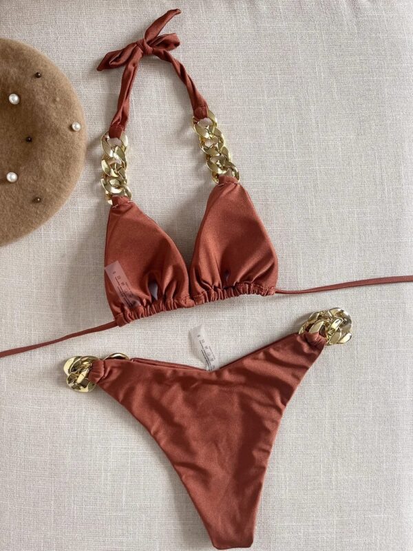 A TWO PIECE BRAZILIAN BIKINI SET with gold chain accents is laid flat on a light fabric surface. A brown beret with pearl decorations is positioned nearby.