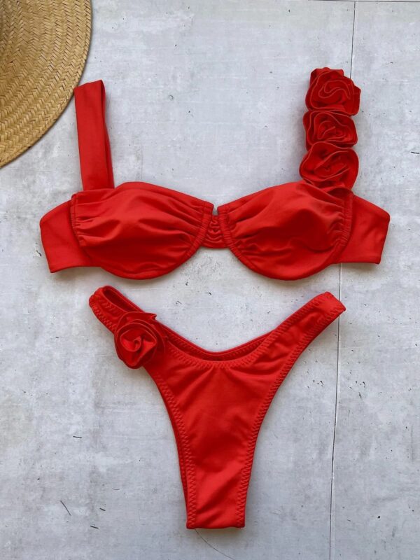 A red FLOWER DIVA TWO PIECE SWIMSUIT with rose embellishments on one shoulder strap and one side of the bottoms, laid out on a light-colored surface next to a straw hat.