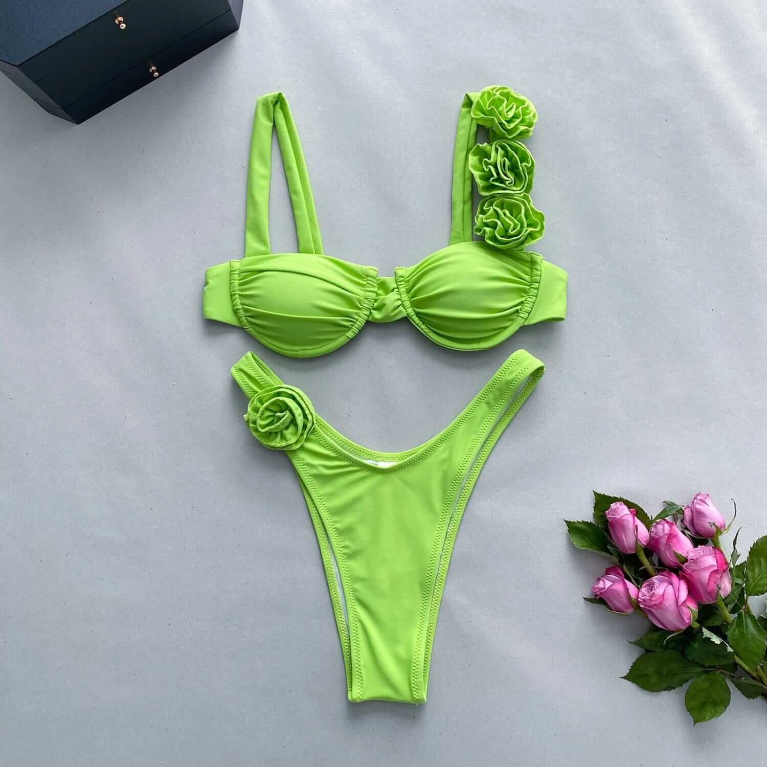 A neon green FLOWER DIVA TWO PIECE SWIMSUIT with floral decorations is displayed on a flat surface next to pink roses and a small, closed black box.