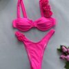 The FLOWER DIVA TWO PIECE SWIMSUIT features a bright pink bikini with an underwire top and high-cut bottoms, adorned with fabric roses on one shoulder strap and one side of the bottoms. The set is elegantly displayed on a light gray surface next to pink roses.