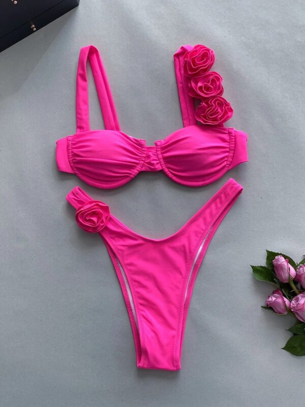 The FLOWER DIVA TWO PIECE SWIMSUIT features a bright pink bikini with an underwire top and high-cut bottoms, adorned with fabric roses on one shoulder strap and one side of the bottoms. The set is elegantly displayed on a light gray surface next to pink roses.