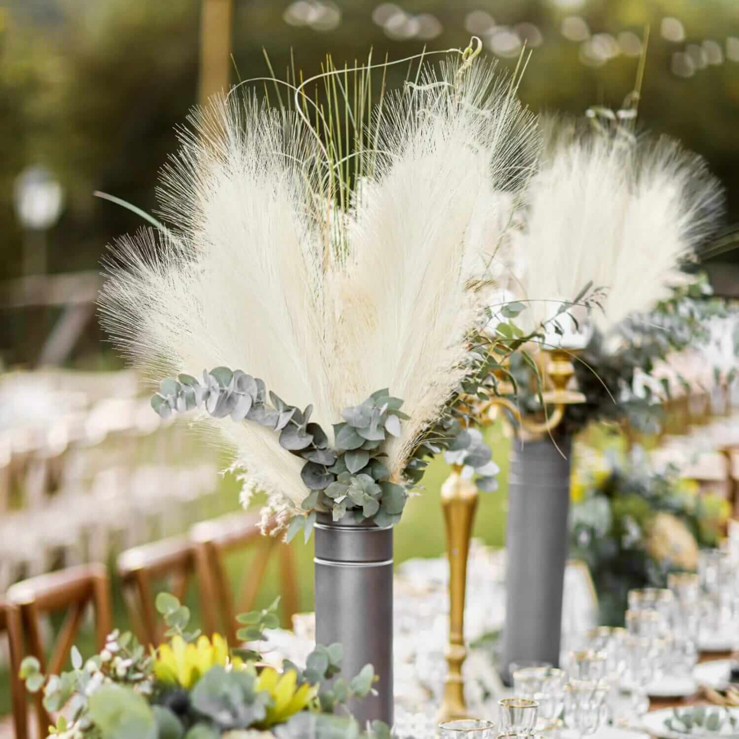 A table setting featuring tall gray vases holding arrangements of Fluffy Pampas Grass and greenery. The setup includes rows of chairs and glasses, suggesting a formal outdoor event.