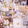 A decorative wedding display featuring cupcakes on a tiered stand, a white cake adorned with 12Pcs Fashion 3D Hollow Butterflies, floral arrangements, candles, and an array of butterflies in gold, silver, and white.