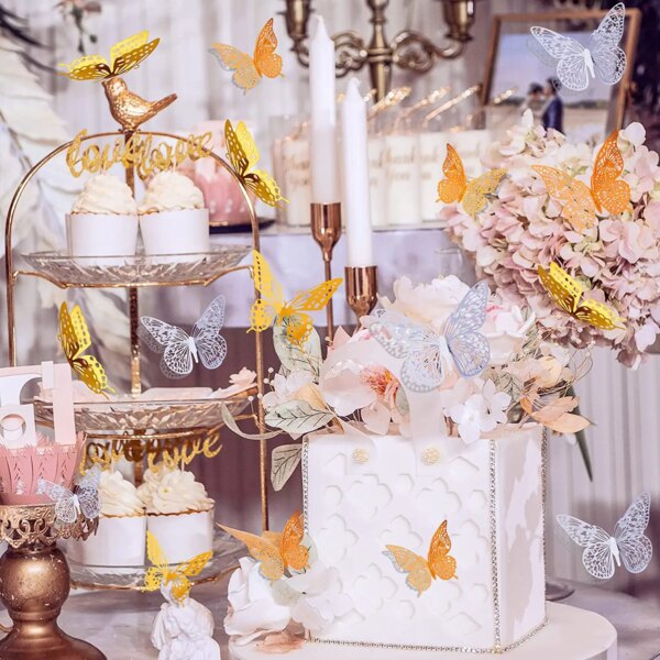 A decorative wedding display featuring cupcakes on a tiered stand, a white cake adorned with 12Pcs Fashion 3D Hollow Butterflies, floral arrangements, candles, and an array of butterflies in gold, silver, and white.