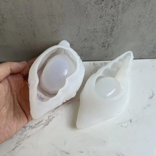 Two white silicone molds, shaped like seashells, are displayed: one is being held and the other is placed on a marble surface against a gray wall. Ideal for crafting your own Seashell Scented Candle Jar.