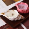 Two shell-shaped candles rest on a wooden tray. One candle is white with gold flakes, and the other is pale pink. Both candles are lit, emitting a beautiful glow reminiscent of the Seashell Scented Candle Jar.