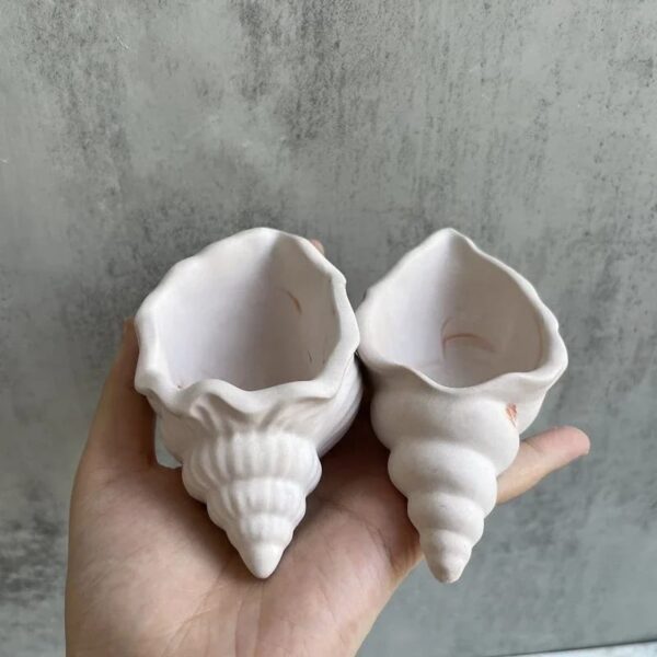 A hand holds two Seashell Scented Candle Jars against a grey background, showcasing their elegant, white, conch-shaped ceramic design.