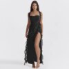 A woman in the Ruffles Trim Square Neck Slit Maxi Dress stands on a white background. The black, sleeveless dress features a square neck and ruffles trim that accentuates the elegance of its thigh-high slit design. She is wearing sandals and has her hands at her sides.