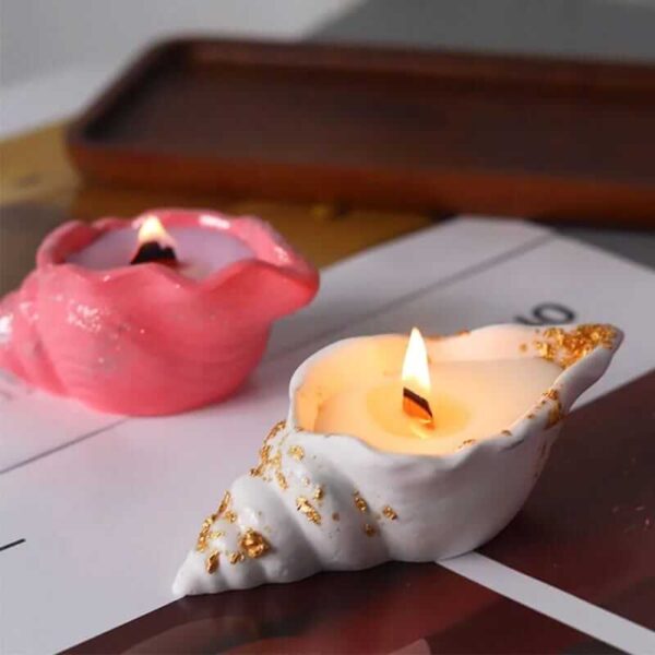 Two lit Seashell Scented Candle Jars, one white with gold accents and the other pink, placed on a table. A brown tray is in the background.