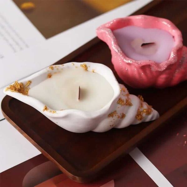 Two seashell-shaped candles, one white with gold accents and one pink, sit on a rectangular wooden tray. The tray is placed on a partially visible surface with paper underneath. A beautiful Seashell Scented Candle Jar complements the coastal ambiance.